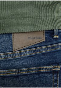 Tissu côtelé vert en haut, jean en denim en dessous. Un patch en cuir beige avec "CHASIN'" embossé, cousu sur le jean. Couture jaune visible.