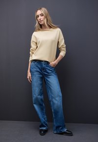 Beżowa bluza z szerokim dekoltem, zestawiona z luźnymi niebieskimi dżinsami. Model stoi na tle ciemnoszarego tła, ma na sobie czarne buty.