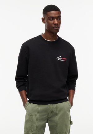 Noor mees taskutes, seljas must Tommy Jeans dressipluus ja helerohelised cargo-püksid, vaadates küljele valgel taustal.