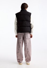 Černá prošívaná vesta s vysokým límcem, nošená přes světlé tričko s dlouhým rukávem, doplněná světle růžovými volnými džínami a béžovými botami.