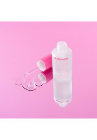 Przezroczysta butelka oleju do włosów z różową nakrętką, otoczona różowym tłem i błyszczącą plamą cieczy, z eleganckim designem.