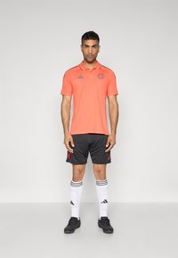 Polo orange avec des accents gris, associé à un short noir avec des logos rouges, des chaussettes rayées blanches et des chaussures de sport noires.