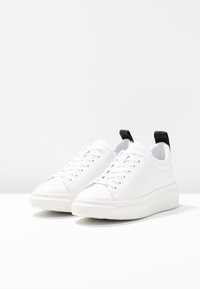 Białe sneakersy o gładkiej strukturze, okrągłym czubku i sznurowanym designie. Posiadają kontrastującą czarną zakładkę na pięcie oraz grubą białą podeszwę.