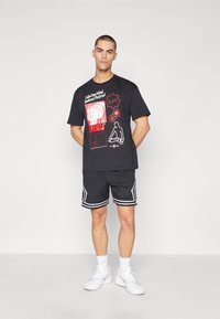 Camiseta gráfica negra con diseños en rojo y blanco, combinada con pantalones cortos de malla negra con ribetes blancos y zapatillas deportivas.