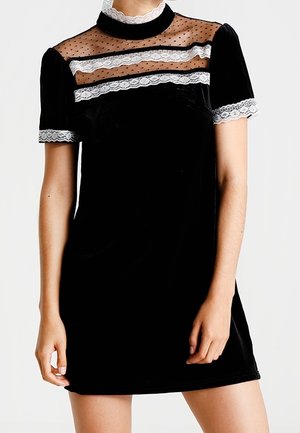 Zwarte fluwelen jurk met korte mouwen, doorschijnende polkadot inzet, witte kanten afwerking bij de halslijn, mouwen en borstdetail.