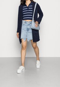 Marinblå texturerad kofta över en randig marinblå och vit kort polo, kombinerad med en slitna jeanskjol och vita platåskor. Bär en ljusblå väska.