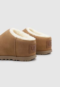 Pantofole marroni in suede con interno in shearling bianco e suola in gomma marrone, dotate di una patch con logo sul tallone.