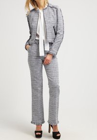 Kvinna i en grå tweedfransjacka och byxor med en vit blus och svarta högklackade platåskor, stående med ena handen i fickan.