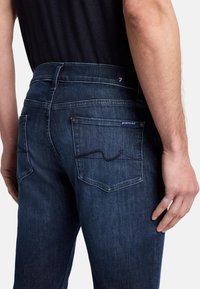 Jeans in denim blu scuro con una vista ravvicinata del retro. Presentano due tasche posteriori, dettagli di cucitura discreti e un'etichetta visibile sulla vita.