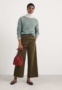 Maglione a maglia verde chiaro, dolcevita beige, pantaloni ampi in corduroy marrone, borsa a secchiello in suede rossa, ballerine nere. Gli accessori includono una cintura e orecchini.