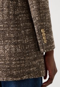Brauner Fischgrät-Blazer aus strukturiertem Stoff, mit einem Detail aus vier Knöpfen an den Ärmeln und im taillierten Schnitt.