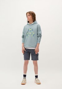 Jasnoniebieska bluza z kapturem z graficznym motywem czaszki w kwiaty, noszona na białej koszulce. Szare szorty cargo, białe i granatowe skarpetki, kremowe sneakersy.
