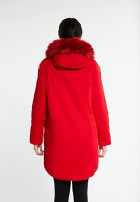 Parka rossa isolata con colletto in pelliccia sintetica, cappuccio, maniche lunghe e orlo leggermente curvo. Tessuto morbido senza chiusure visibili.