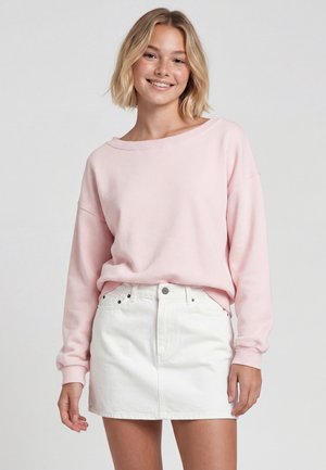 Giovane donna sorridente, indossa una felpa rosa chiaro a maniche lunghe e una minigonna bianca in denim, in piedi davanti a uno sfondo semplice.