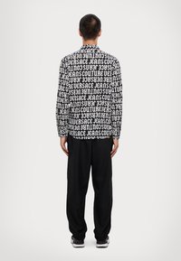 Long-sleeve shirt with black and white repeated text pattern, paired with black trousers. The shirt features a standard collar and button cuffs.