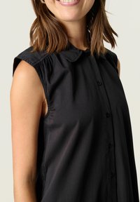 Blusa sin mangas negra con un cierre de botones en el frente, textura suave y un cuello clásico. Presenta un sutil plisado en los hombros.