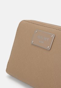 Carteira texturizada bege com uma placa de logotipo prateada, apresentando um fecho com zip e uma forma compacta e retangular.