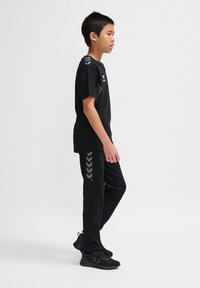 Schwarzes Sportoutfit bestehend aus einem enganliegenden T-Shirt und einer Hose. Die Hose hat graue Chevron-Akzente. Getragen mit schwarzen strukturierten Sneakers.