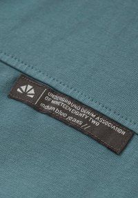 Turkos tyg med ribbad textur, med en svart vävd etikett. Etiketten innehåller texten: "UNDERGROUND DENIM ASSOCIATION OF NINETEEN EIGHTY TWO."