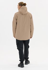 Beige vattentät jacka med huva, med en avslappnad passform och en böjd nederkant, kombinerad med svarta cargobyxor och svarta skor.