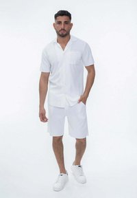 Chemise blanche à manches courtes avec une poche et un short assorti, en tissu léger, associé à des baskets blanches sur un fond uni.