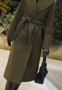 Alyvuogių spalvos paltas su apvyniotu dizainu, odinės detalės ir surišama dirželis. Derinamas su juodomis pirštinėmis ir struktūrizuota rankine.