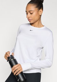 Balts garām piedurknēm sporta tops ar melnu Nike logotipu, kopā ar matēti melnu ūdens pudeli. Gluda tekstūra, pieguloša forma, mūsdienīgs siluets.