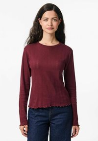 Tamsiai raudonas ilgomis rankovėmis viršus su tekstūruotu briaunų raštu ir volangais, derinamas su tamsiai mėlynomis džinsinėmis kelnėmis. Glotnus audinio paviršius.