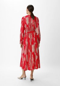 Femme aux cheveux bruns en queue de cheval basse portant une robe midi à manches longues rouge et rose clair avec des motifs abstraits et des escarpins beiges, de dos.