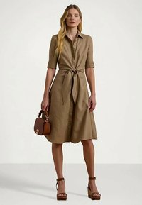 Γυναίκα φορώντας καφέ κοντό φόρεμα με ζώνη μπροστά, κρατώντας καφέ τσάντα, και φορώντας καφέ πλατφόρμες, στέκεται μπροστά από απλό φόντο.