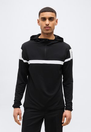 Mladý muž s krátkymi vlasmi, oblečený v čiernej mikine s bielym horizontálnym pruhom cez hruď a horné paže, stojaci na bielom pozadí.