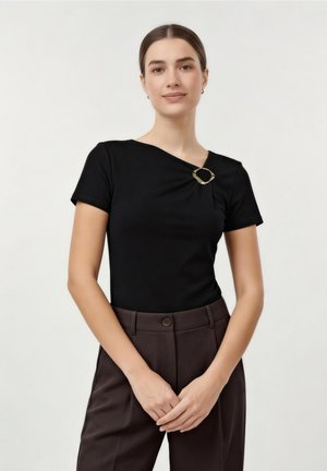 Donna che indossa una maglietta nera a maniche corte con una spilla d'oro sul colletto e pantaloni scuri a vita alta, in piedi con le mani intrecciate.