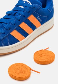 Sapatilhas de camurça azul com riscas laranjas, apresentando cadarços azuis e uma sola de borracha branca. Duas rolos de cadarços laranjas estão posicionados abaixo.