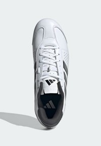 Bílá adidas teniska s černými pruhy, šněrování uvázané, s polstrovaným límcem, pohled zhora na světlém šedém povrchu.