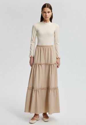 Ženska z ravnimi rjavimi lasmi nosi kremno dolg rokav in bež večplastno dolgo krilo z volančki, stoji pred enobarvno podlago.