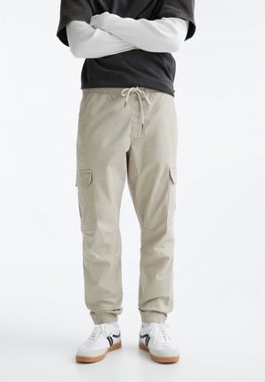 Personne portant un pantalon de jogging cargo beige avec taille à cordon, poignets élastiques, baskets blanches, t-shirt noir à manches courtes sur un t-shirt blanc à manches longues.