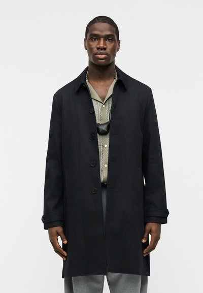 Άνδρας που φοράει ένα μαύρο παλτό με κουμπιά πάνω από ένα εμπριμέ ανοιχτοπράσινο πουκάμισο, ανοιχτόγκρι παντελόνι και μια μαύρη θήκη λαιμού, στέκεται μπροστά σε ένα απλό φόντο.