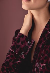 Alyvuogių aksominis marškiniai su juodu leopardų raštu, turintys rankogalius su sidabro užsegimais ir subtiliu blizgesiu, arti nufotografuota, kad būtų rodomi detalės.