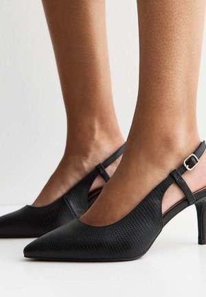 Zwarte gestructureerde slingback hakken met spitse neuzen, gedragen op blote voeten tegen een effen lichte achtergrond.