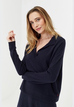 Donkerblauwe pullover met een kraaghals, ribgebreide manchetten en zoom. Zachte textuur met een comfortabele pasvorm. Kleine rode accentdetail aan de zijkant.