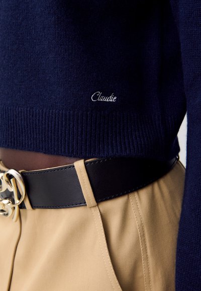 Pull en maille marine avec ourlet côtelé, portant le mot "Claudie" brodé en blanc. Associé à une ceinture en tissu beige et en cuir noir.