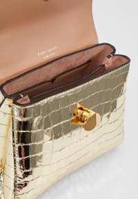 Złota torebka Kate Spade z tłoczeniem w zjawiskowy krokodyl, z otwartą klapą, złotym zapięciem na obrót i jasnobrązowymi skórzanymi przegródkami wewnątrz.