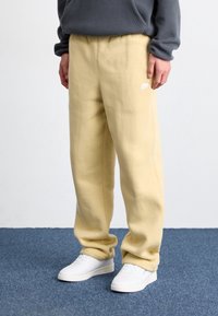 Person klädd i beigefärgade Nike joggingsbyxor och vita sneakers står på en blå matta med en grå sweatshirt delvis synlig.