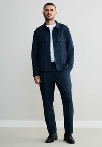 Navy button-up jacket with front pockets, paired with matching trousers and a white t-shirt, complemented by black shoes. Smooth texture.