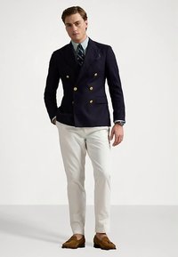 Tengerészkék, dupla soros aranygombos blézer világoskék inggel és fehér nadrággal párosítva. A megjelenést barna mokaszin teszi teljessé.