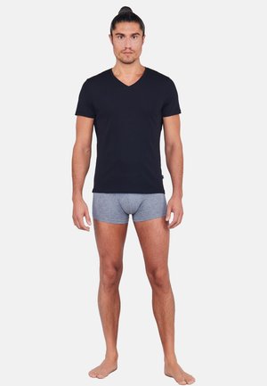 Mann mit nach hinten gebundenem Haar, der ein schwarzes V-Ausschnitt-T-Shirt und graue Boxershorts trägt, steht barfuß vor einem schlichten Hintergrund.