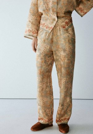 Pantalon beige et orange à motif floral avec poignets de chemise assortis, porté avec des chaussures en daim marron sans lacets, debout devant un fond clair.