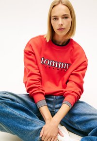 Červená mikina s logom "tommy" v bielej a čiernej farbe, manžety s rebrovaním s čierno-bielym pruhom, doplnená o voľné modré džínsy z denimu.