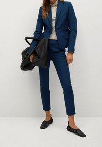 Costume bleu marine sur mesure avec un blazer cintré et un pantalon court, associé à un col roulé clair et des ballerines noires texturées. Sac fourre-tout noir.