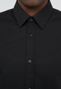 Chemise noire boutonnée avec col, présentant une texture lisse et six boutons noirs assortis sur le devant.